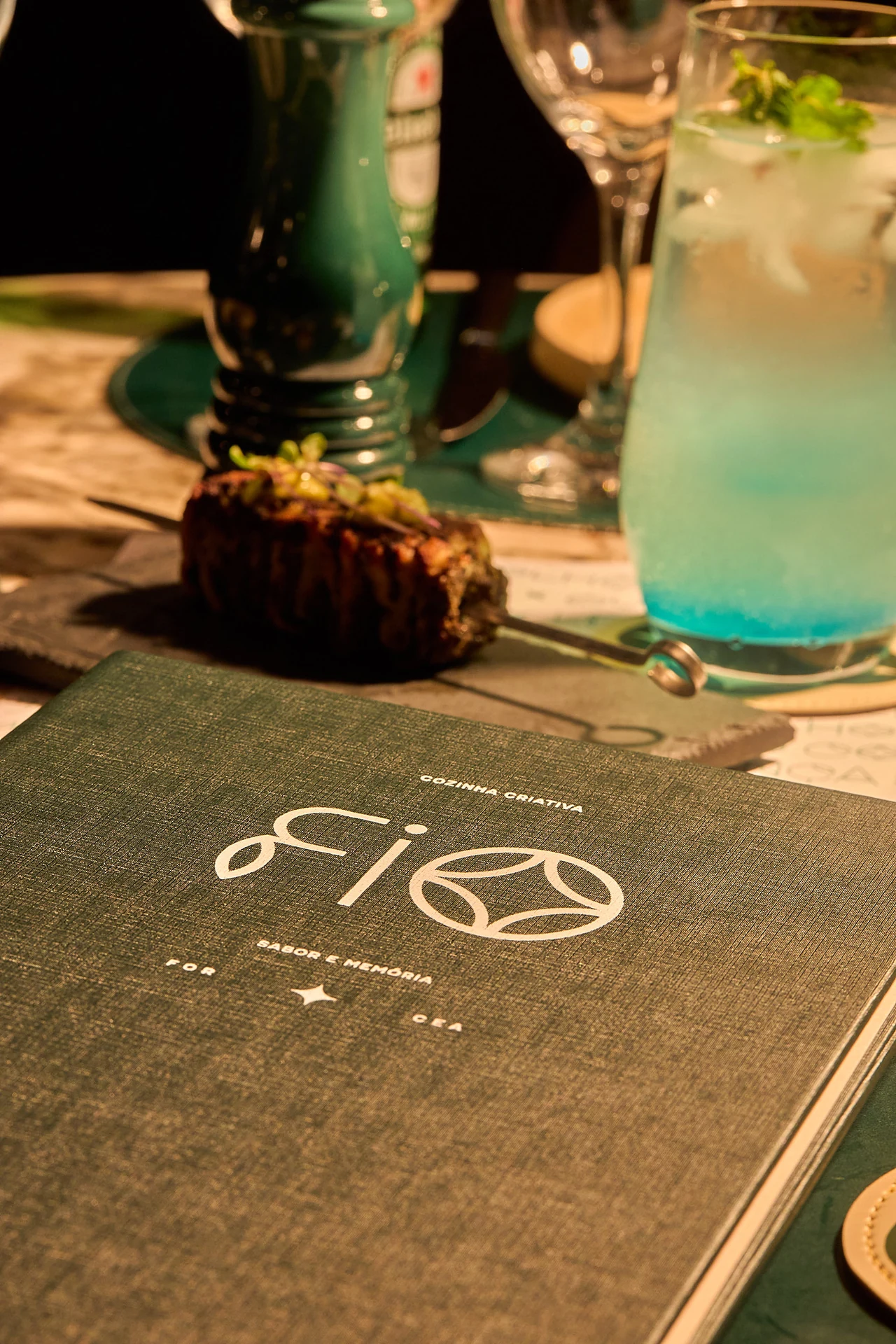 Mesa do Fio com o cardápio, um drink autoral e um prato da casa